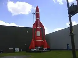 A model of a rocket-shaped spacecraft with three engine nacelles around its cylindrical main body. Photographed standing upright in front of a building.