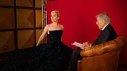 A photograph of Bennett sitting in an armchair sketching a portrait of Gaga, who is stood beside him waring a black dress.