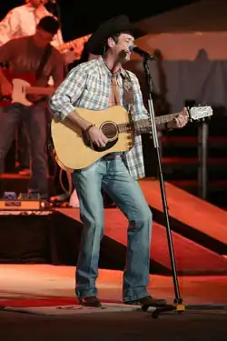 A man wearing a black cowboy hat, a checked shirt and jeans, playing guitar and singing into a microphone