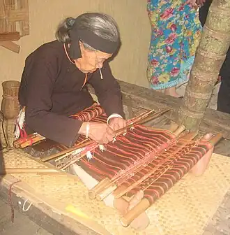This Hlai weaver tensions her traditional backstrap loom with her feet. She is using a large number of slim heddle rods, attached to only a few warp threads; these are sometimes called pattern rods. Hainan Island, Southern People's Republic of China.