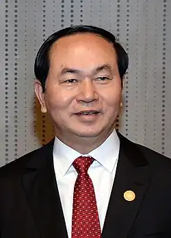 Trần Đại Quang looking a bit to the right, wearing a black suit, white shirt and a red tie while having a red-yellow pin on his right chest.