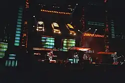 An elaborate concert stage set, set in a dark stadium. Three automobiles hang above two projector screens, aiming their headlights towards the stage.