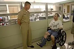 Color photograph of a standing man in uniform (left) and man in wheelchair with casts on right foreleg and left forearm (right).