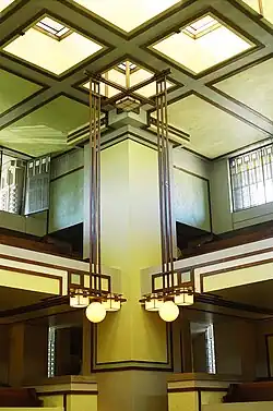 One of the square piers at the corner of Unity Temple's auditorium. The ceiling has square skylights with wooden boards at their borders. Hanging from the ceiling are chandeliers with globes and cubes.