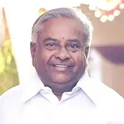 V. Muniyappa wearing a white shirt, standing and smiling at a public event