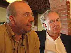 An image of Michele Santoro (and Vauro Senesi) in a room with one wearing brown clothing and the other a white shirt and black coat