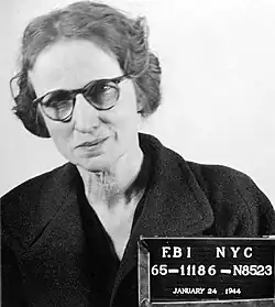 A grayscale mugshot of a leaning white woman from the chest-up; she has medium-dark hair, and is wearing a dark coat and spectacles. Included is a placard that says, "F.BI NYC, 65-11186-N8523, January 24 1944".