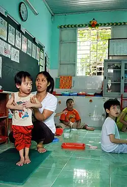 A woman is helping a young boy to stand up in a classroom with other students