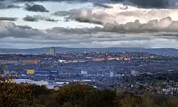 Trees in the foreground; houses, shops and industrial buildings in the middle ground; and hills in the background. The sky is grey.