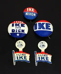 Photograph of five buttons on a black background. The first round button has "IKE" in white on red and "NIXON" in white on blue. The second round button has "I LIKE" in white on red, "IKE AND DICK" in blue on white and a white five-pointed star on blue. The third round button has a red band, a white band with "IKE" in blue and a blue band. The fourth and fifth buttons are shapes as waving flags, with a red band, a white band with "I LIKE IKE" in blue and a blue band. Above the flag a round piece has the logo of a lithographers' union from Chicago.