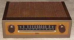 A small rectangular brown box with a metallic coppery top on a beige carpet. On its front are two dials and a tuner with a range between 88 and 108