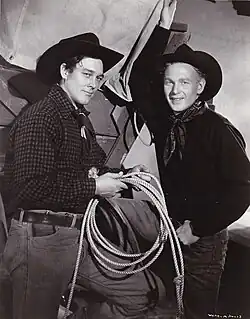 Black and white photograph showing two men standing. They are dressed in cowboy garb: jeans, kerchiefs around their necks, and cowboy hats. The man on the left is holding a lasso near his waist. A section of what appears to be a covered wagon is visible behind them.