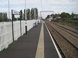 A photo taken from a railway platform to the left of two tracks. There is no platform to the right and a sign on the platform reads "Watlington". Overhead gantries are visible above the line and a train is visible in the background on the left-hand track. There is a level crossing and signal box before the right-hand platform in the distance behind that.