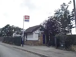 A tiny brick building with a shuttered over window and a closed blue door. A path passes to the left and to the left of that there is a pole with a double arrow sign, an IntaLink logo, and a sign that reads "Watton-at-Stone". A sign above above the window and door on the building also reads "Watton-at-Stone".