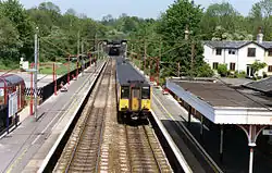 A small train is on the right-hand side of two parallel tracks which are flanked by station platforms, covered with station canopies. At the back, the tracks disappear into a tunnel mouth below a hill