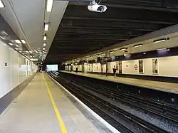 Several people walking inside a building with a black ceiling and white walls bordered in black and displaying several advertisements