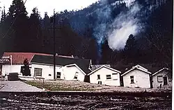 A color photograph of shifted homes and smoke in the background following the flood