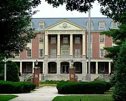 A brick neoclassical building fronted by four light-toned columns, with a green lawn and sidewalk in the foreground