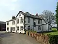 A pathway with a wall on the right leads to a white-painted two-storey house with attic and black-painted dressings; a lower two-storey wing extends to the left and on the right is a lean-to extension