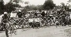 A group of around ten cyclists, climbing over a fence. Behind the fence are cars.