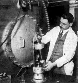 Old photograph of a man with a moustache and a lab coat, holding up a complicated piece of equipment