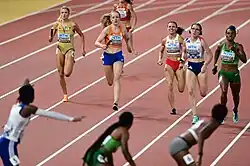 Photo of (six visible) runners on red athletics track seen from the front approaching (three visible) runners of the next heat seen from the back