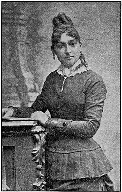 A young Black woman with hair in an elaborate updo, wearing a corseted dress or suit with a pleated peplum, standing in a portrait studio