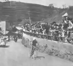 Black-and-white photograph of a cyclist on a road with spectators on a wall next to the road