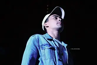 Young JV is pictured wearing a denim jacket and a baseball cap