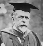 A black and white photo of an older white man with a beard in academic robes and a graduation cap