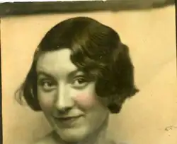 Black and white photo of a woman sitting in a boat with an oar in her hands. She smiles into the camera and has light skin and dark hair.